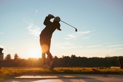 Young junior golfer practicing in a driving range with beautiful sunset light in winter.