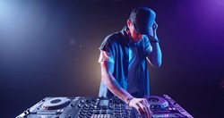 Cool hipster disc jockey performing in a nightclub at a mixer controller, spotted by colorful lights on smoked black background - nightlife concept 
