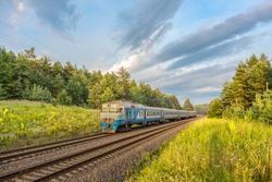 An old broken diesel passenger train rides a suburban route on rails through the forest in the rays of the setting summer sun. Rural hinterland. Russian railways