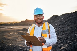 A young Black African coal mine foreman looking into camera smiling wearing reflective bib and hard hat is inspecting samples of coal on his clipboard after a long day of work on site at the coal mine