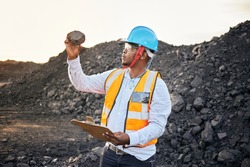 A young Black African coal mine foreman wearing reflective bib and hard hat is inspecting samples of coal after a long day of work on site at the coal mine