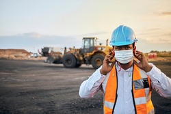 A young Black African coal mine foreman wearing reflective bib and hard hat is putting on a covid 19 face mask after a long day of work on site at the coal mine