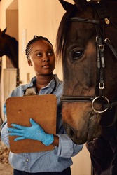 A young Black African woman veterinarian standing outside a horse stable holding a clipboard after checking the health of a horse. 