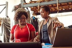 Two black African Zimbabwean ladies working in a textile factory with the supervisor helping employee with measurement using laptop computer measuring tape and steel ruler