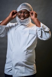 Young black African male chef putting on a hair net in studio wearing a chefs jacket black toque hat and beard net while smiling and laughing