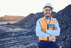 A young African mine worker wearing protective wear is looking at the camera with coal mine in the background