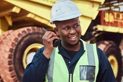 A young African mine worker is talking on a two way radio in front of a large haul dump truck wearing his personal protective wear