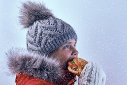 A girl in outerwear is eating a burger. Child eats street food in heavy snow outdoors.
