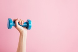 Womans hand holding blue dumbbell isolated on pink background. Equipment for home workout. Fitness and activity. Sport and healthy lifestyle concept. Copy space in right side