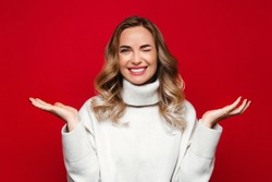 Charming woman with happy surprise on her face spreading her hands to the side, smiling and winking, wear white sweater, isolated on red background, copy space. Winter season and sale