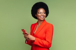 Successful african businesswoman hold smartphone look aside with confident happy smile. Afro american young entrepreneur female in luxury red suit with mobile phone isolated over green studio wall