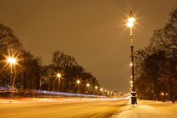 a snow-covered avenue with burning lanterns on a winter evening. along the road there are black trees against the sky