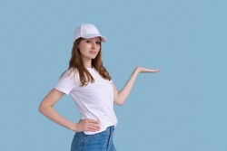 Young happy surprised smiling female promoter 20s in white casual t-shirt and cap pointing with hand to the side at workspace copy space mock up isolated on blue background studio portrait.