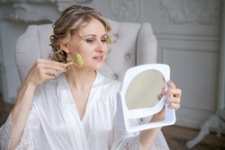 Beautiful woman in silk robe using jade face roller with natural stones doing massage holding mirror in hand while sitting in armchair doing morning routine. Skin care beauty treatment concept at home