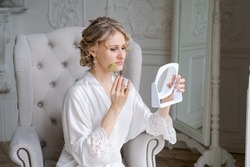 Beautiful woman in silk robe using jade face roller with natural stones doing massage holding mirror in hand while sitting in armchair doing morning routine. Skin care beauty treatment concept at home