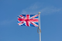 Union Jack flag on flagpole in strong breeze, flying to the left, left side is facing camera, blue sky with small cloud.