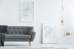 Elegant, gray sofa with wooden legs and large map posters on a white wall in a designer minimalist living room interior of an architect