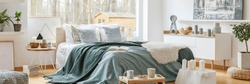 Green blanket on a cozy, double bed by a large window in a scandinavian style bedroom interior with a view at a forest