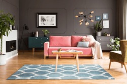 Wooden table on blue carpet in grey living room interior with fireplace and pink sofa. Real photo
