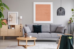 Rose gold painting and poster in bright loft interior with grey sofa near cabinet and wooden table. Real photo