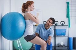 Smiling professional personal trainer helping sportswoman exercising with ball