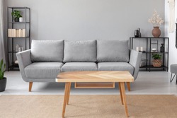 Wooden table on carpet in front of grey sofa in minimal living room interior with plant. Real photo