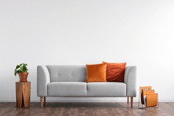 Comfortable couch with orange and red pillow in spacious living room interior, real photo with copy space on the empty white wall