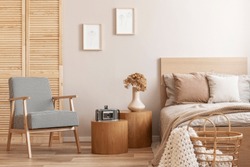 Trendy retro armchair next to two round wooden tables with vintage camera and vase with flowers in beige bedroom interior