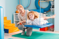 Woman helping a smiling boy to exercise on a therapy swing