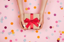 Woman hands holding present box with red bow on pastel pink background with multicolored confetti. Flat lay style.