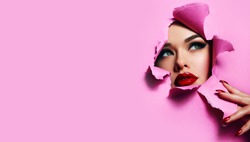 the face of a young beautiful girl with a bright make-up and with plump red lips peeks into a hole in pink paper. Nails with bright red lacquer.Lipstick,cosmetics,makeup, fashion, beauty,beauty salon.
