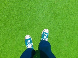 Man wearing old green sneakers in blue trousers standing on green grass outdoor.