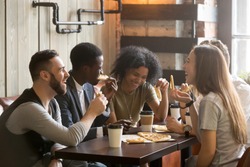 Multiracial happy young people eating pizza in pizzeria, black and white cheerful mates laughing enjoying meal having fun sitting together at restaurant table, diverse friends share lunch at meeting