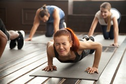 Young fit sporty woman with painful face expression doing hard difficult plank fitness exercise or push press ups feeling pain in muscles at diverse group training class in gym, endurance concept