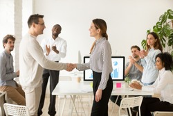 Smiling female boss promoting rewarding handshaking motivated worker showing respect while team applauding congratulating colleague at group meeting, appreciation and employee recognition concept