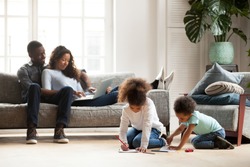 Black family spend free time together in living room at new home. Married couple sitting on couch use computer, little daughter and son have a fun on a warm wooden floor drawing with colourful pencils