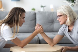 Old mother and grown millennial daughter in law arm wrestling having fight or family conflict, young and senior women rivals competing, disagreement and different generations problems concept