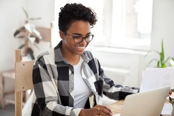 Smiling black woman sit at desk working at laptop review paperwork documents, happy African American female using computer managing financial papers, girl in glasses studying or chatting on pc