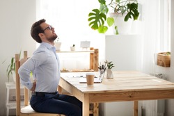 Tired millennial office worker stretch in chair suffer from sitting long in incorrect posture, male employee have back pain or spinal spasm working in uncomfortable position. Sedentary life concept