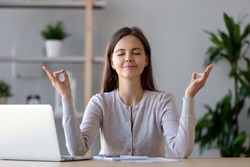 Calm young woman worker taking break doing yoga exercise at workplace, happy mindful female student meditating at home office desk feel balance harmony relaxation, stress relief zen at work concept