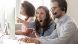 Female call center agent operator telemarketer helping male colleague teaching new worker online customer support pointing at computer, business team in wireless headset talk working together on pc