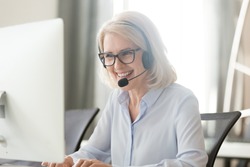 Happy old businesswoman in headset speaking by conference call looking at computer, mature female aged call center agent operator telemarketer talking consulting customer service support in office