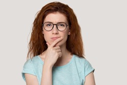 Pensive young red-haired young woman isolated on grey background look at camera thinking, thoughtful redhead girl in glasses pondering planning problem solution, lost in thoughts consider idea