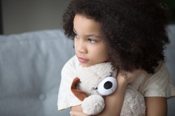 Close up upset stray biracial kid girl sitting on sofa cuddle stuffed toy dog looks aside feel lonely unhappy. Preschool age orphan, bullying discrimination, depression, psychological trauma concept