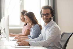 Smiling call center male worker pose looking at camera girls colleagues on background, sales agent wearing headset use pc answers incoming phone calls, provide professional support to customer concept