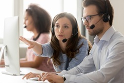 Call center employees wearing headset using computer, woman explains show to new colleague marketing database program, busy service phone operators sitting at shared desk, assistance teamwork concept