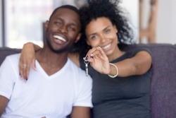 Excited African American young family show keys to own home, happy black couple sit on couch praise buying first house together, smiling husband and wife purchase new property. Ownership concept