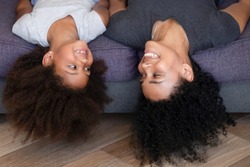 Happy young African American mom and small daughter have fun together lying upside down on couch, excited kid play with smiling nanny or sister, enjoy spending time together fooling around at home