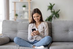 Smiling teenager girl relax sit on sofa at home using cellphone texting chatting with friend, happy young woman on couch hold smartphone shop online or check mobile application or social media