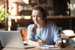 Focused woman wearing headphones using laptop in cafe, writing notes, attractive female student learning language, watching online webinar, listening audio course, e-learning education concept